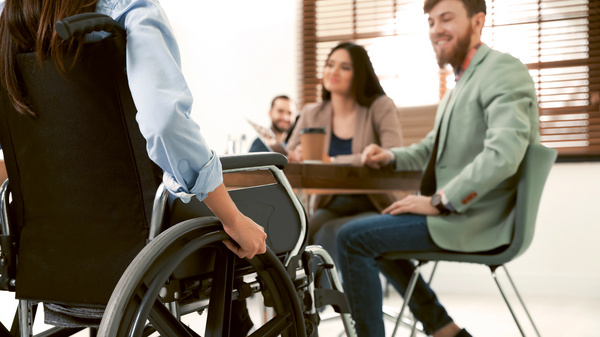 Dargestellt ist eine Szene mit einer Person im Rollstuhl, die auf einen Tisch zurollt, an dem zwei nicht eingeschränkte Personen sitzen.