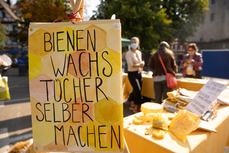 Zu sehen ist ein Plakat, auf dem in Großbuchstaben steht: "Bienenwachstücher selber machen". Im Hintergrund ist ein Tisch mit Utensilien erkennbar.