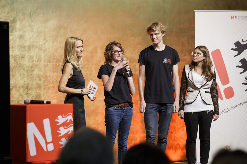 Drei junge Menschen stehen auf der Bühne neben der Moderatorin. Die Jugendbeirätin hält das Mikrofon und spricht hinein.