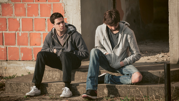 Dargestellt sind zwei junge Männer, die missmutig im Freien nebeneinander sitzen.
