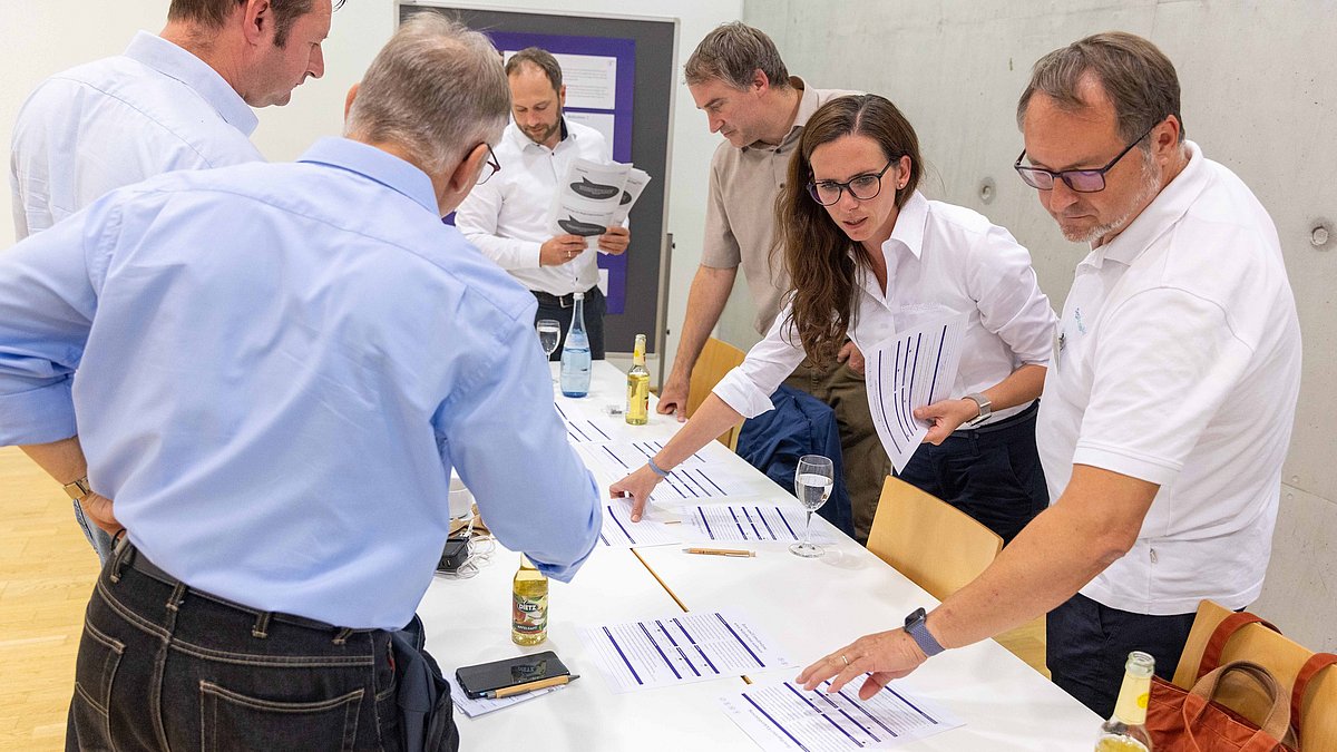 Workshop-Teilnehmende stehen um einen Tisch, eine Frau hat DINA4-Papier in der Hand.