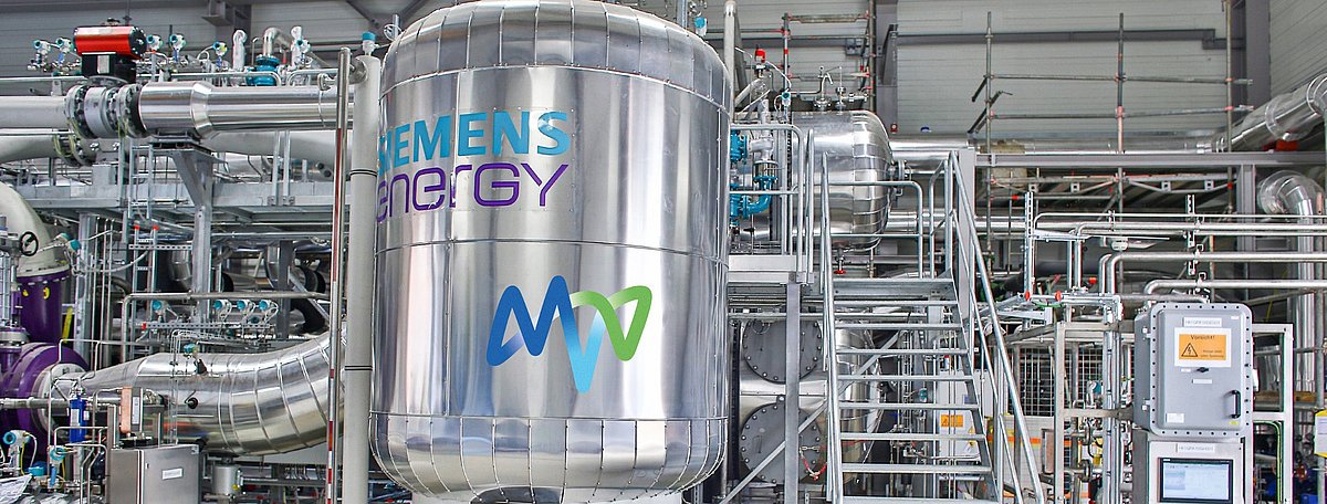 Farbfoto einer großen, silbernen Anlage mit der Aufschrift „Siemens Energy“. Viele Rohre, Leitungen und eine Metalltreppe sind sichtbar.