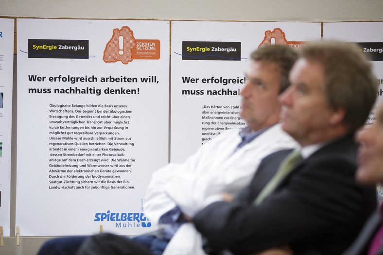 Nachhaltigkeitstage 2014 Minister Untersteller neben einem Mann im weißen Kittel, sitzend. Im Hintergrund Plakate.