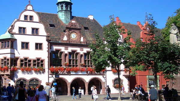 Ein Platz in einer Kleinstadt mit historischen Gebäuden. Menschen flanieren über dem Platz und sitzen im Schatten unter Bäumen.