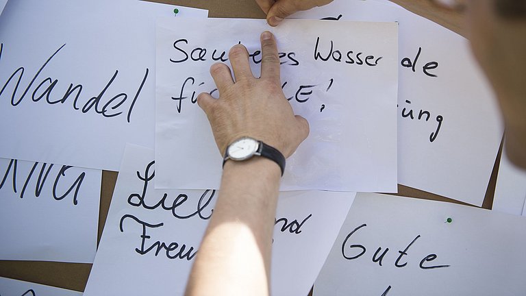 An einer Pinnwand sind weiße Papierstücke mit verschiedenen Aufschriften angeheftet. Ein Mann pinnt einen Zettel mit der Aufschrift : Sauberes Wasser für Alle! an die Pinnwand.