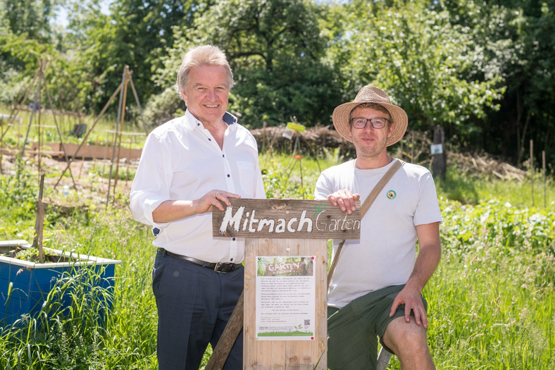 Minister Franz Untersteller posiert für ein Foto in einem Garten mit einem Mann, auf einem Schild steht "Mitmach Garten"