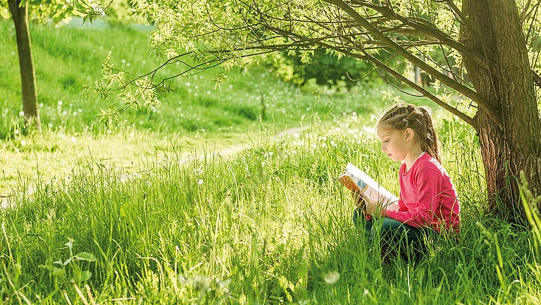 Ein Mädchen sitzt unter einem Baum auf einer Wiese und liest in einem Heft.