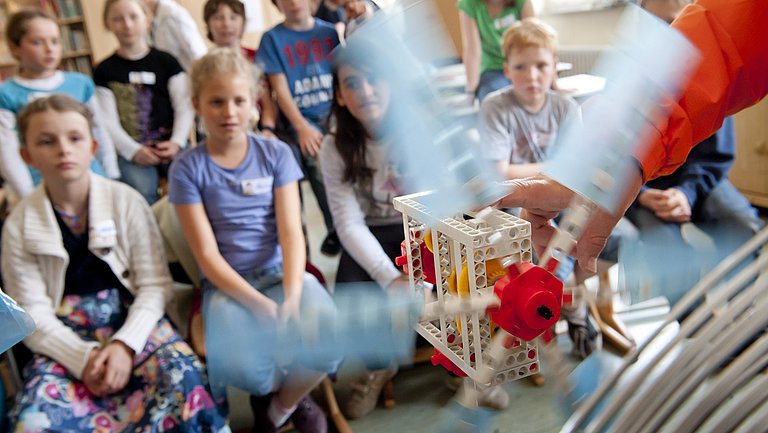 Kinder sitzen im Klassenzimmer. Im Vordergrund sieht man, dass ein Modell, eine Art Propeller bedient wird.