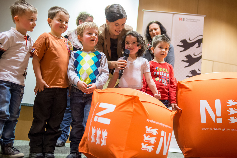 Fünf Kinder im Kindergartenalter stehen auf der Bühne. Die Moderatorin der Preisverleihung beugt sich von hinten zu einem Mädchen und hält das Mikrofon in der Hand.