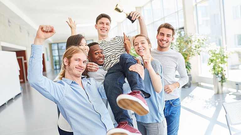 Fünf Jugendliche, die freudig einen sechsten Jugendlichen hochheben. Alle lächeln in die Kamera und strecken einen Arm in die Höhe. Der sechste Jugendliche hält einen kleinen Pokal in der linken Hand.  