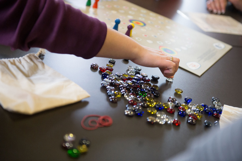 Ein Arm ragt ins Bild. Eine Spielsituation auf einem Tisch. Bunte Glassteine liegen auf dem Tisch, in Hintergrund ein Spielbrett. Ein Stoffbeutel.