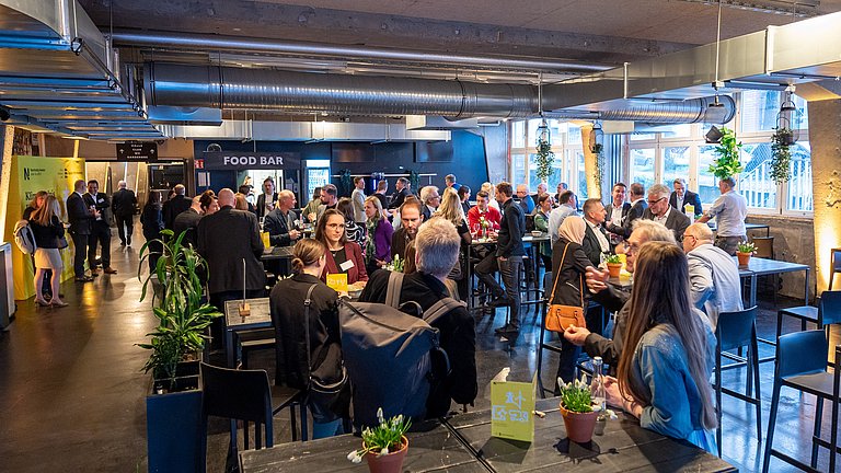 Man sieht viele Menschen stehend an Bartischen. Sie unterhalten sich und netzwerken.