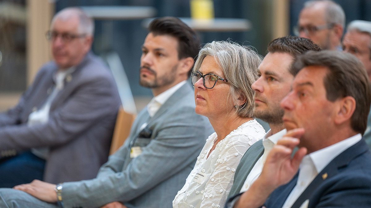 Farbige Fotografie: Eine Gruppe von Personen sitzt in einer Reihe und hört aufmerksam zu. Im Fokus eine Frau mit Brille in weißer Bluse.