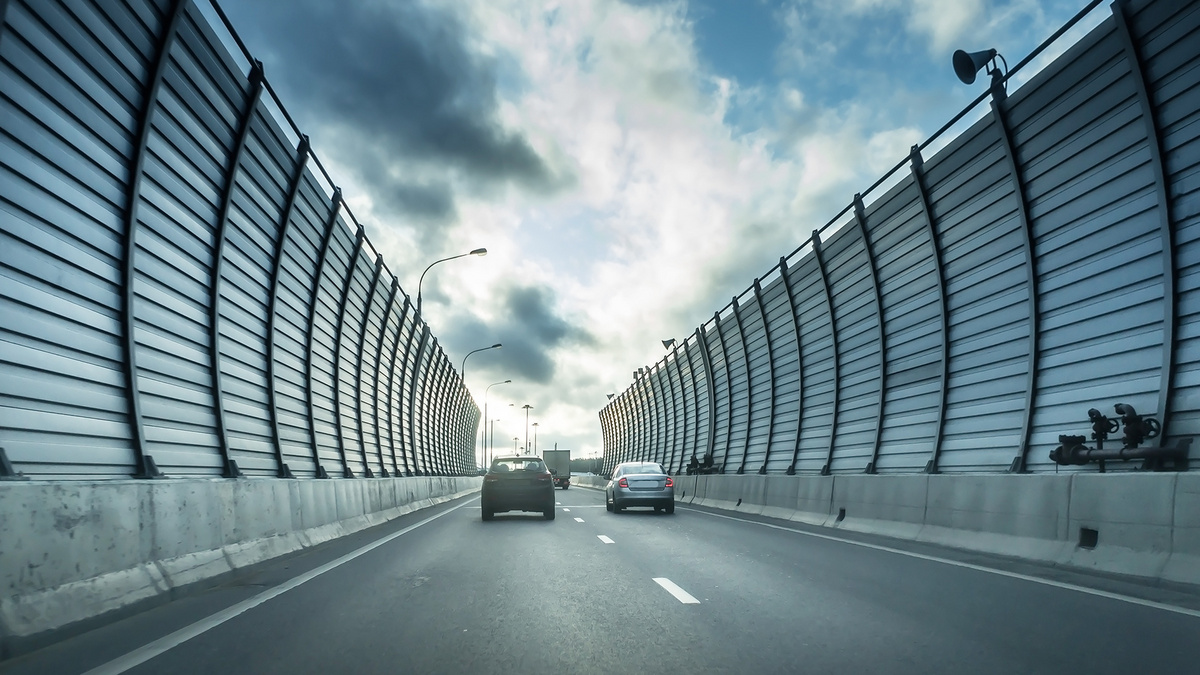 Autos fahren auf einer Straße, die mit hohen Lärmschutzwänden ausgestattet sind. Da der Blick der Straße folgt, ergibt sich ein Tunneleindruck. Über dem Stückchen Himmel, das die Lärmschutzwände noch freigeben, treiben dicke Wolken.