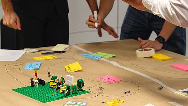 Ein Tisch mit Kleinlego-Bauteilen und Männchen und Beschriftungen auf Papier, darum herum stehen Menschen und arbeiten an dem Projekt.