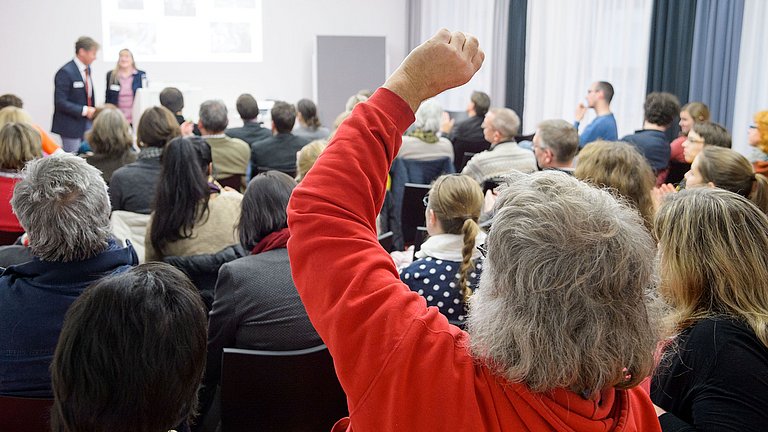 Lehrerinnen und Lehrer sitzen in einem Vortragsraum. Sie hören zwei Vortragenden zu oder melden sich für eine Wortbeitrag.