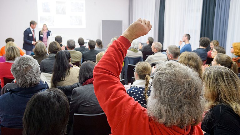 Lehrerinnen und Lehrer sitzen in einem Vortragsraum. Sie hören zwei Vortragenden zu oder melden sich für eine Wortbeitrag.