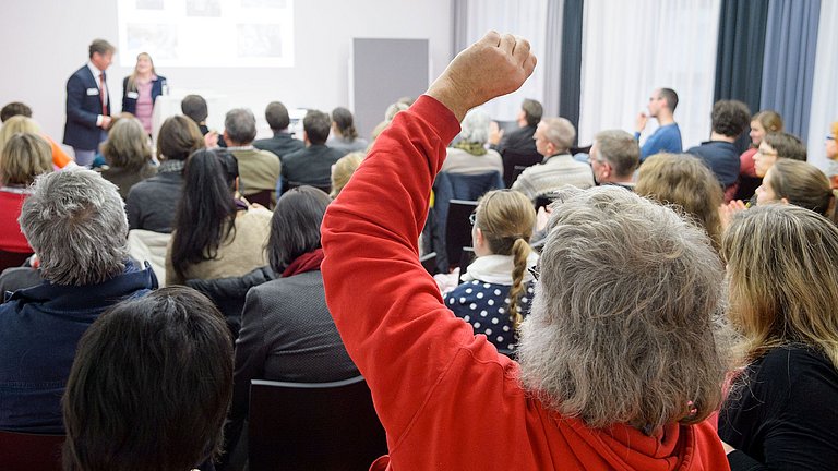Lehrerinnen und Lehrer sitzen in einem Vortragsraum. Sie hören zwei Vortragenden zu oder melden sich für eine Wortbeitrag.