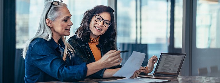 Zwei Geschäftsfrauen im Gespräch an einem Arbeitstisch mit Laptop und Dokumenten.