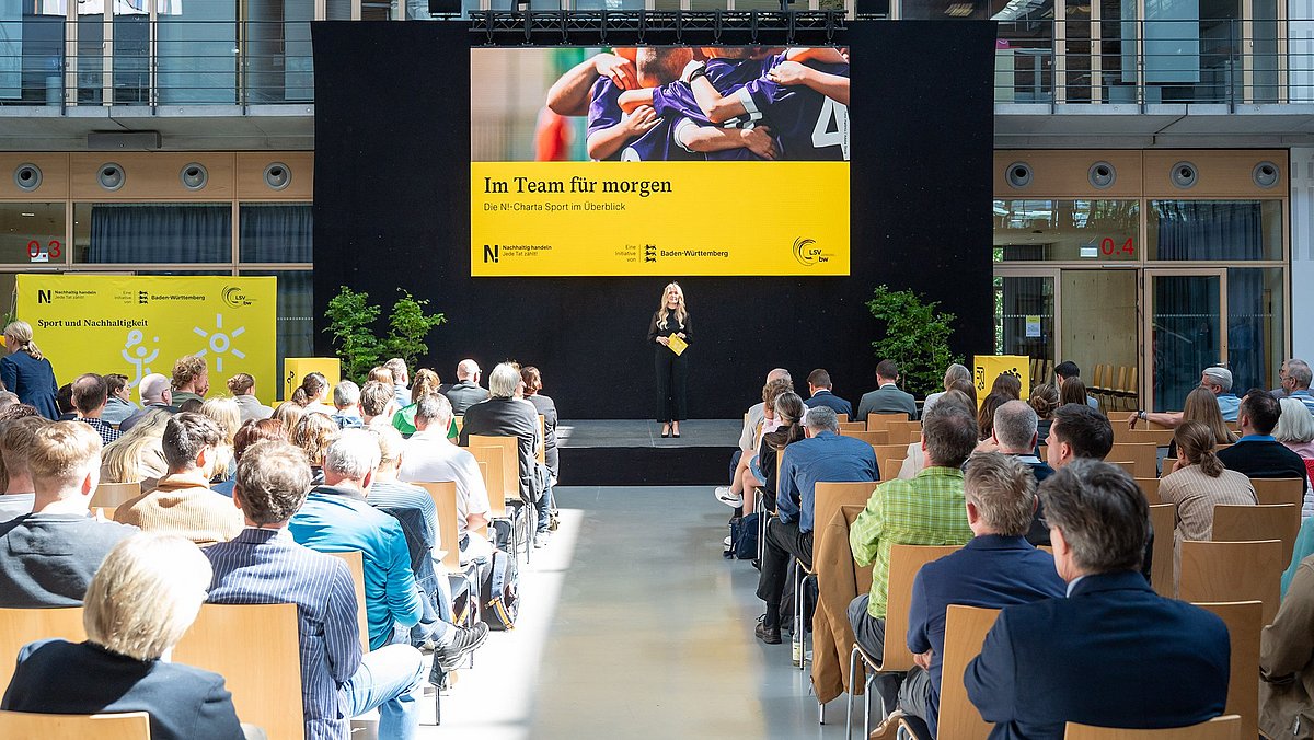 Farbige Fotografie: Publikum blickt auf eine Bühne mit großer Leinwand. Dort steht eine Frau mit Mikrofon vor dem Text 'Im Team für morgen – Die N!-Charta Sport im Überblick'.