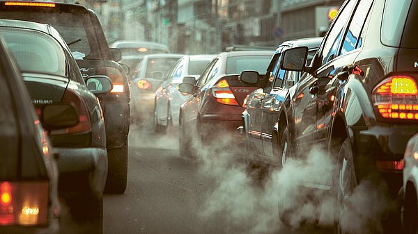 Dargestellt sind Autos im Straßenverkehr. Abgase treten deutlich sichtbar aus den Auspuffrohren heraus.