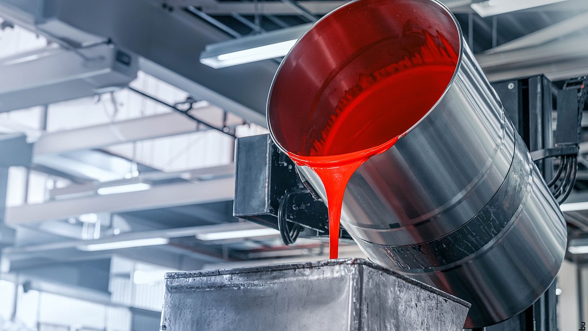 In einer hellen Produktionshalle fließt aus einem großen Metallfass rote Farbe in den Container einer Abfüllanlage.