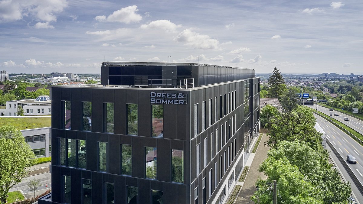 Kastenförmiges Bürogebäude mit großen Fenstern und der Aufschrift "Drees & Sommer", daneben eine Straße und im Hintergrund Bäume und Häuser.