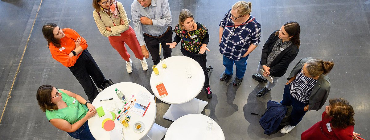 Von oben wird ein Blick auf eine Gruppe von Bildungsakteuren geworfen, die um drei Stehtische herumstehen und ein Thema erarbeiten.