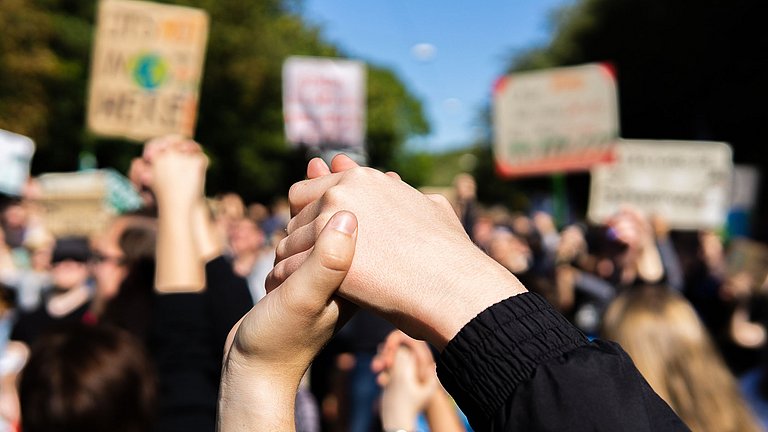 Im Vordergrund sind zwei ineinander umschlungene Hände zu sehen, die in die Höhe gehalten werden. Im Hintergrund erkennt man eine Gruppe von Menschen, die beschriftete Plakate in die Höhe halten.