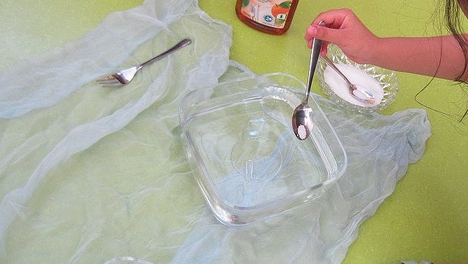 Ein Gefäß mit Wasser, daneben eine Falsch Spülmittel und eine Kinderhand, die einen Löffel zum Wasser hält.