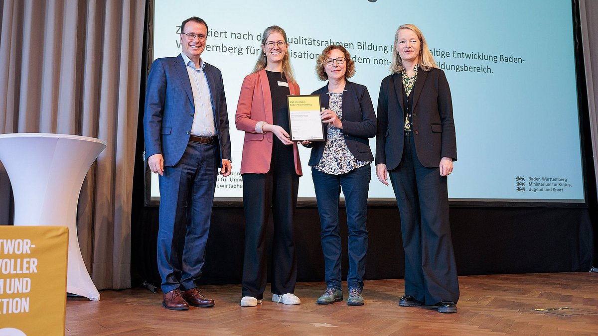 Umweltministerin Thekla Walker MdL und Kultusstaatssekretär Volker Schebesta MdL stehen zusammen mit zwei Frauen auf einer Bühne. Sie lächeln alle in die Kamera, die zwei Frauen halten ein eingerahmtes BNE-Zertifikat.