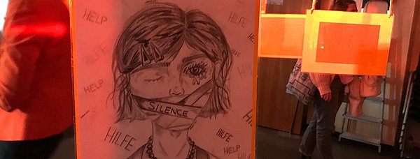 Das Farbfoto zeigt eine Ausstellungsszene mit einer Zeichnung, die ein Mädchen mit einem Pflaster über dem Mund zeigt, darauf steht "Silence". Worte wie „Hilfe“ und "Help" sind um sie herum skizziert.