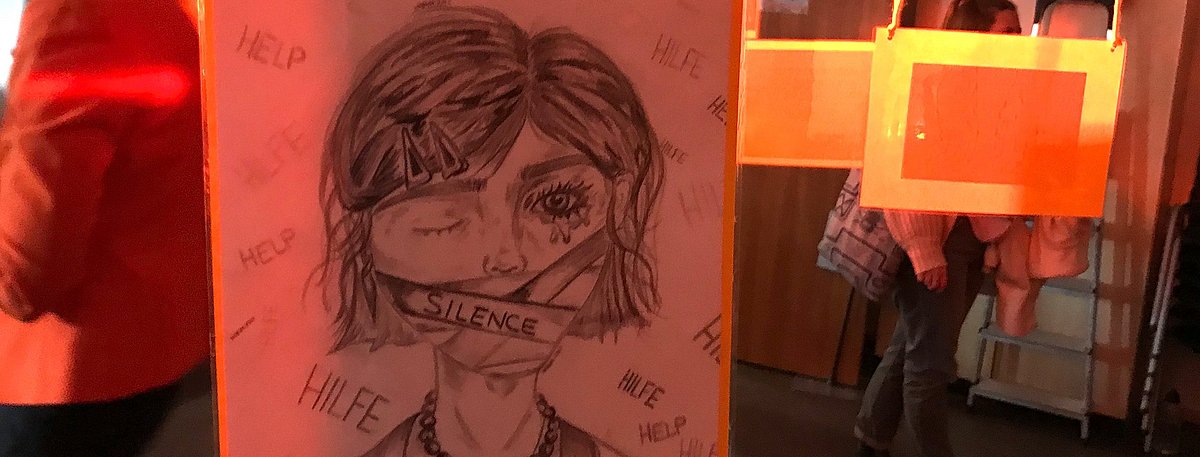 Das Farbfoto zeigt eine Ausstellungsszene mit einer Zeichnung, die ein Mädchen mit einem Pflaster über dem Mund zeigt, darauf steht "Silence". Worte wie „Hilfe“ und "Help" sind um sie herum skizziert.