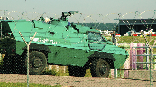 Ein gepanzertes, grünes Bundespolizeifahrzeug steht hinter einem Stacheldrahtzaun und bewacht einen Flughafen.
