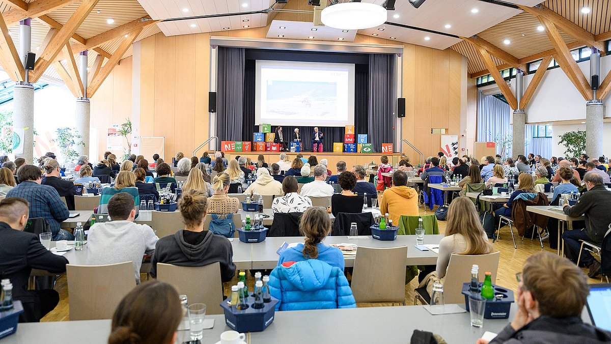 Das Bild zeigt eine Veranstaltungshalle mit Blick auf die Bühne und vielen Personen, die an Tischen sitzen und zur Bühne schauen.