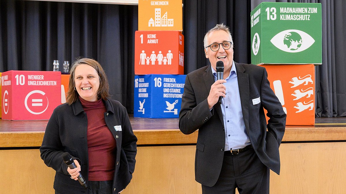 Frau Dengler und Herr Beule stehen mit Mikrophonen in der Hand vor der Bühne und unterhalten sich freudig.