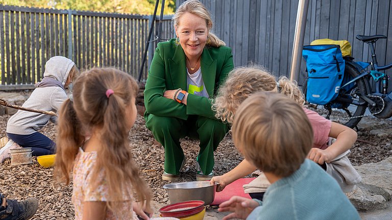 Umweltministerin Thekla Walker sitzt in der Hocke und spricht mit Kindern im Garten.