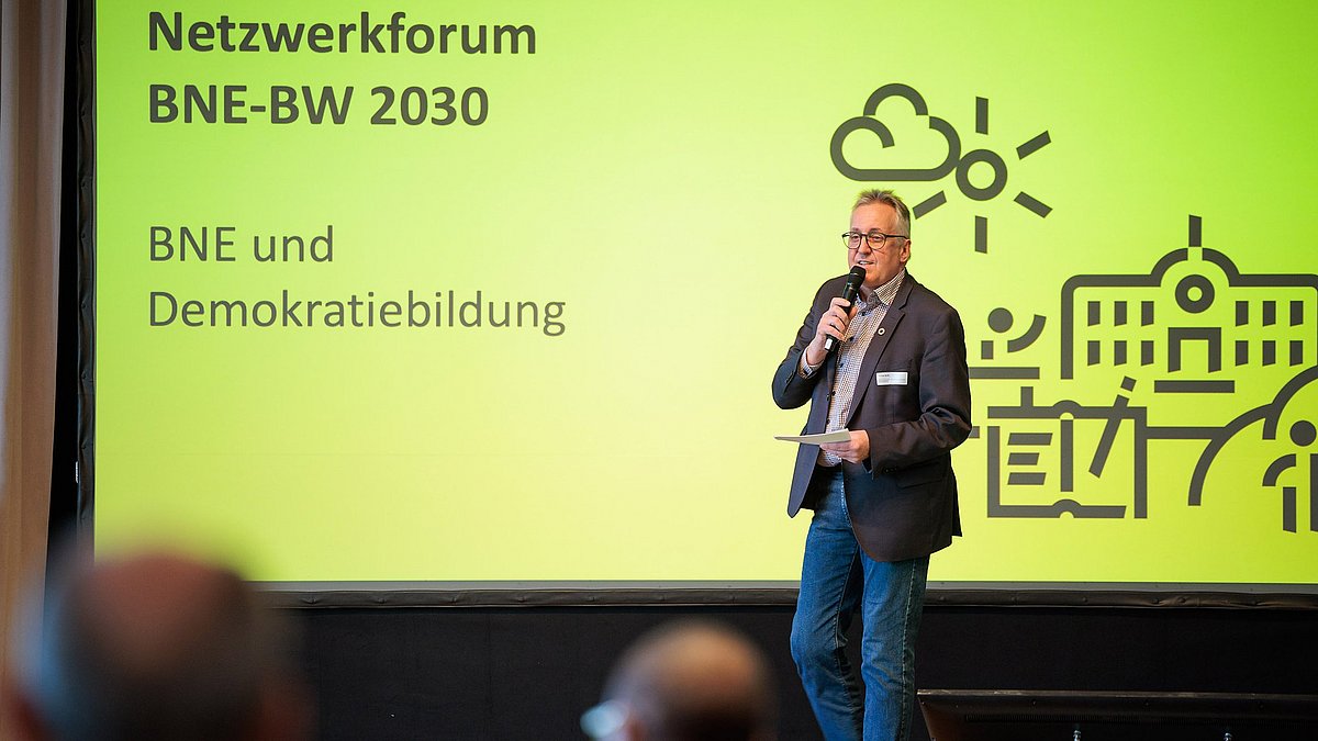 Ein Mann mit einem Mikrofon steht auf einer Bühne. Hinter ihm eine gelbe Präsentationsfolie mit der Aufschrift: „Netzwerkforum BNE-BW 2030 – BNE und Demokratiebildung“