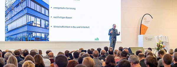 Auf einer Bühne tragt ein Redner vor großem Publikum eine Präsentation zum Thema Zukunft Albau vor.