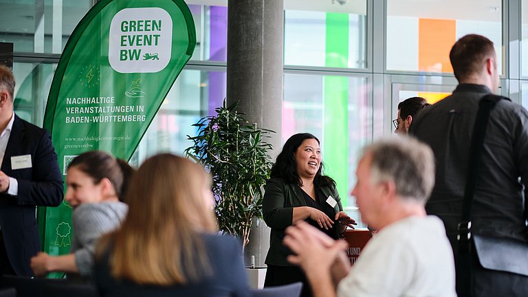Im Vordergrund und Hintergrund sieht man Personen, die sich unterhalten. Im Hintergrund steht ein Aufsteller zum Thema Green Event BW.