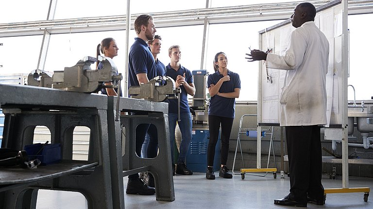 Junge Erwachsene und ein Lehrer in einer Werkstatt. Die Gruppe der Schüler hört dem Lehrer im weißen Kittel zu.