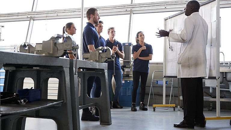 Junge Erwachsene und ein Lehrer in einer Werkstatt. Die Gruppe der Schüler hört dem Lehrer im weißen Kittel zu.
