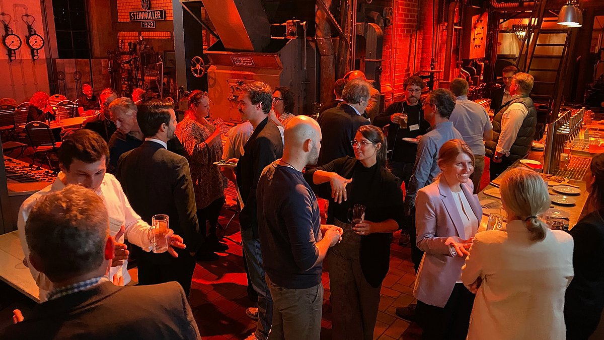 Blick in einen Veranstaltungsraum mit Baratmosphäre. Besucherinnen und Besucher unterhalten sich angeregt.