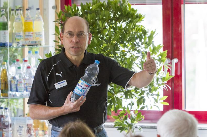 Ein Mann in schwarzen T-Shirt hält eine Mineralwasserflasche in der Hand und spricht. Im Vordergrund sitzendes Publikum.