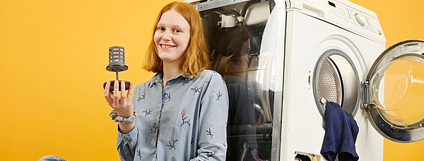 Junge Frau sitzt vor einer Waschmaschine und hält ein Bauteil, orangefarbener Hintergrund, lächelt in die Kamera.