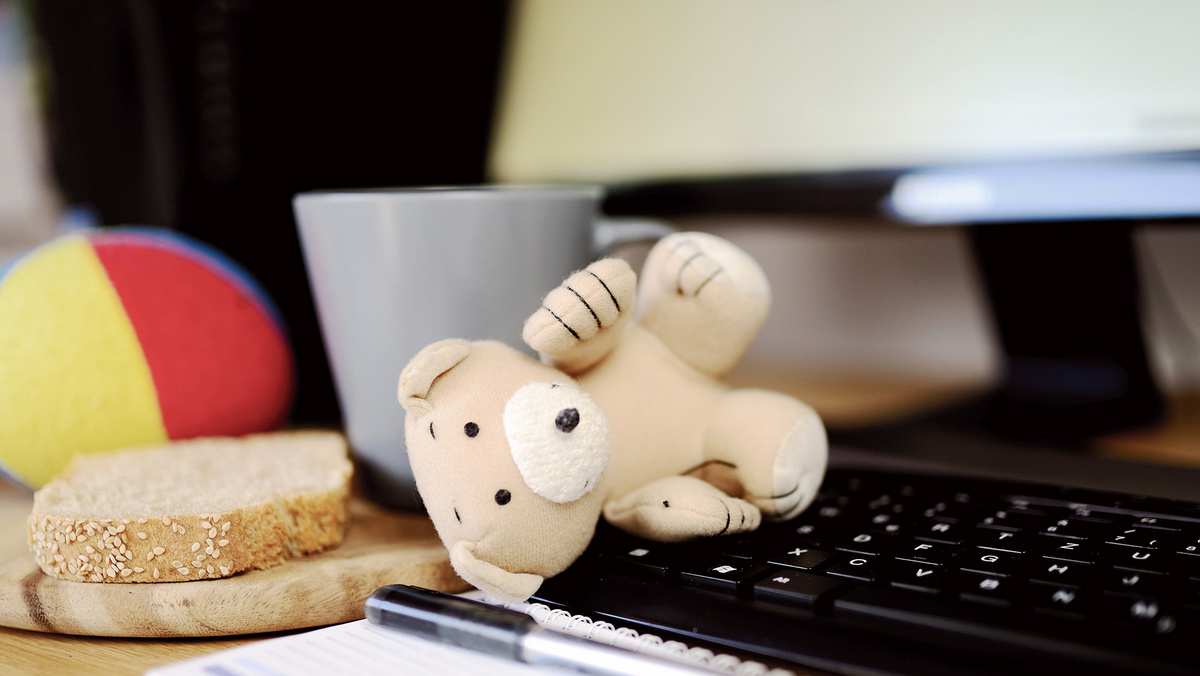 Homeoffice mit Kind Auf einer Computertastatur liegt ein Stofftier, daneben ein Vesperbrett mit einer Scheibe Brot, ein Stoffball und ein Kaffebecher.