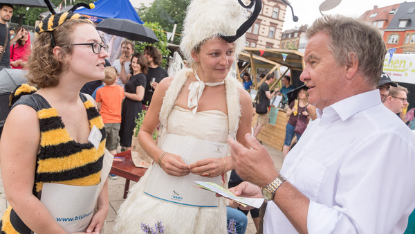 Herr Minister Franz Untersteller unterhält sich mit zwei Frauen, die sich als zwei Insekten verkleidet haben, auf dem Übermorgen Markt