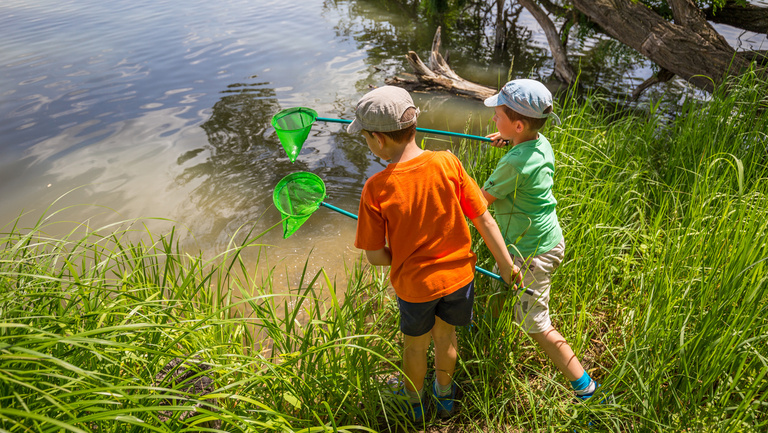 Kinder erleben Wasser Zwei Jungen stehen am Ufer eines Gewässers und wollen mit einem Köcher im Wasser schöfpen.