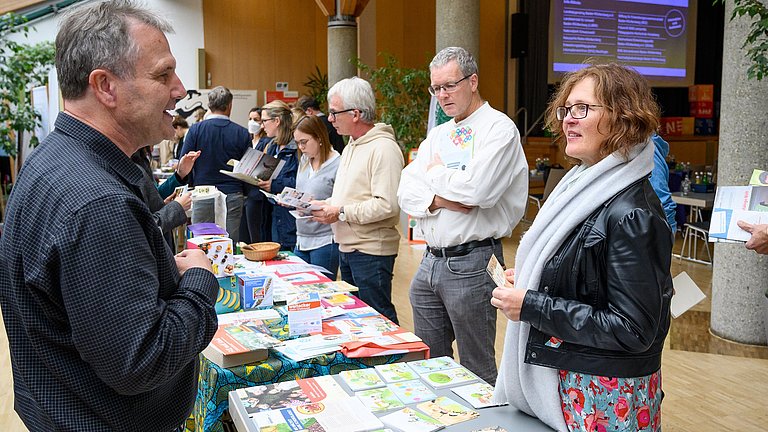 Die Aufnahme zeigt einen Ausschnitt eines Informationsstandes. Links im Bild steht ein Mann hinter dem mit Informationsmaterial ausgelegtem Tisch, der sich mit einer Frau über den Tisch hinweg unterhält. Im Hintergrund sind weitere Personen zu sehen.