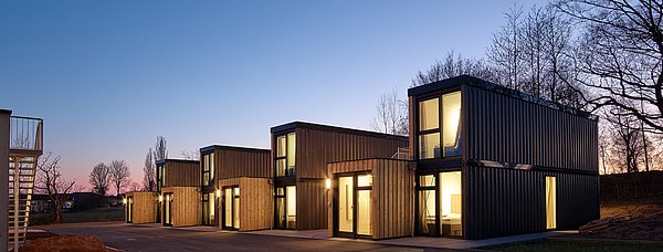 Moderne Wohncontainer aus Holz und Metall mit leuchtenden Fenstern bei Abenddämmerung, umgeben von Bäumen und beleuchtetem Vorplatz.